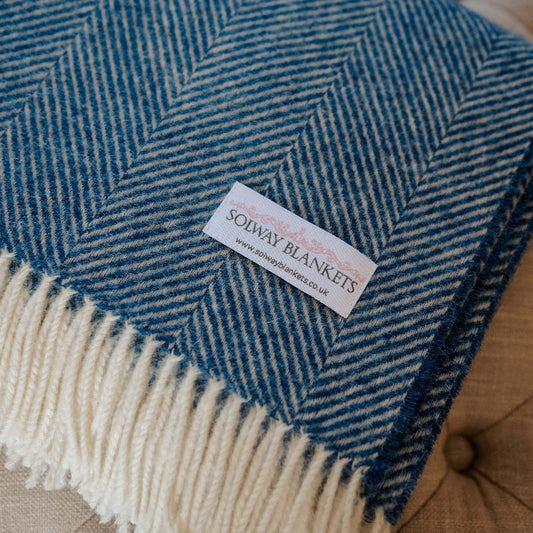 Blueberry Swirl Shetland Wool Herringbone Blanket. Blue and white herringbone patterned blanket with a Solway Blankets woven label on a footstool