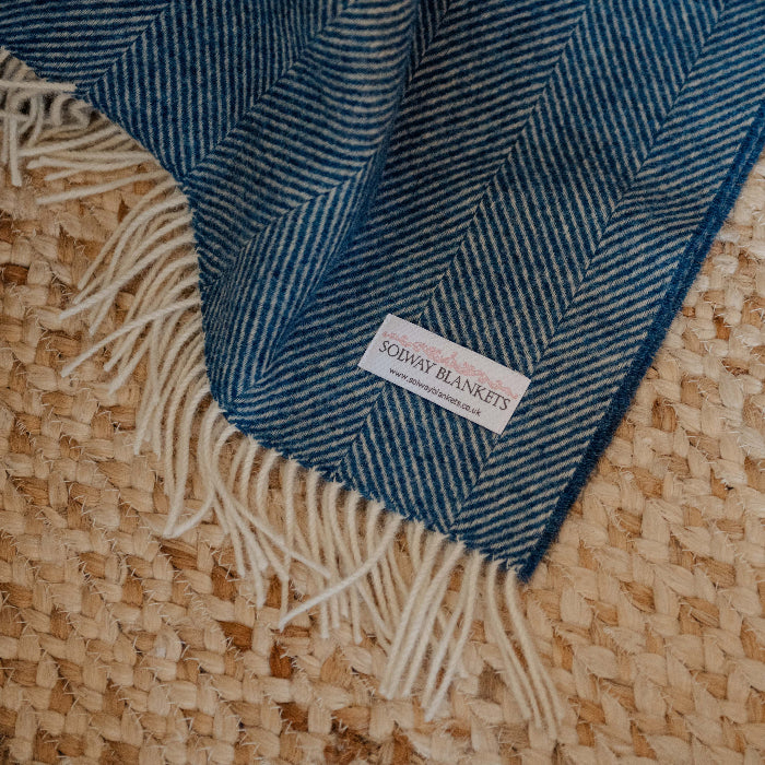 Blueberry Swirl Shetland Wool Herringbone Blanket with white fringe on a textured surface, featuring a brand label.