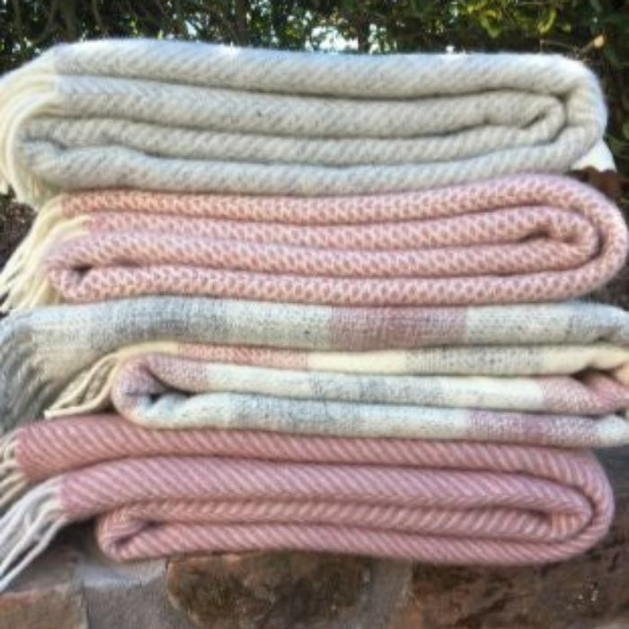 Stack of woolen blankets, one pink herringbone styled, one pink, grey and cream check, one pink honeycomb and one grey herringbone style folded on a stone surface with greenery in the background