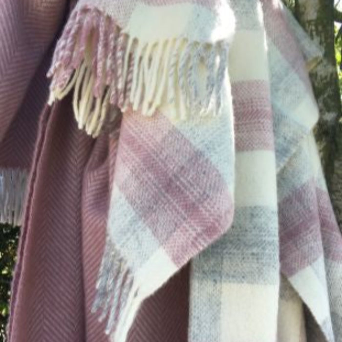 Plaid wool blanket with fringes draped over a tree with colours of pink, cream and grey alongside another pink blanket in a herringbone style. 
