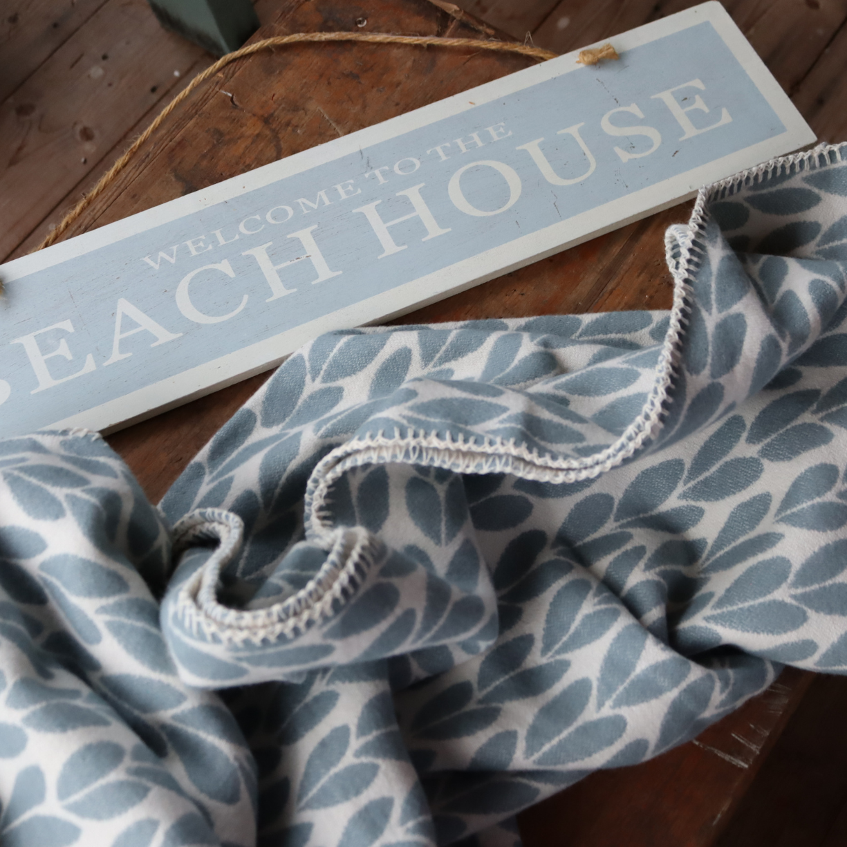 A draped cotton throw with a duck egg blue sycamore leaf pattern on a white background, draped over a rustic wooden surface next to a wooden sign that reads “WELCOME TO THE BEACH HOUSE.”.
