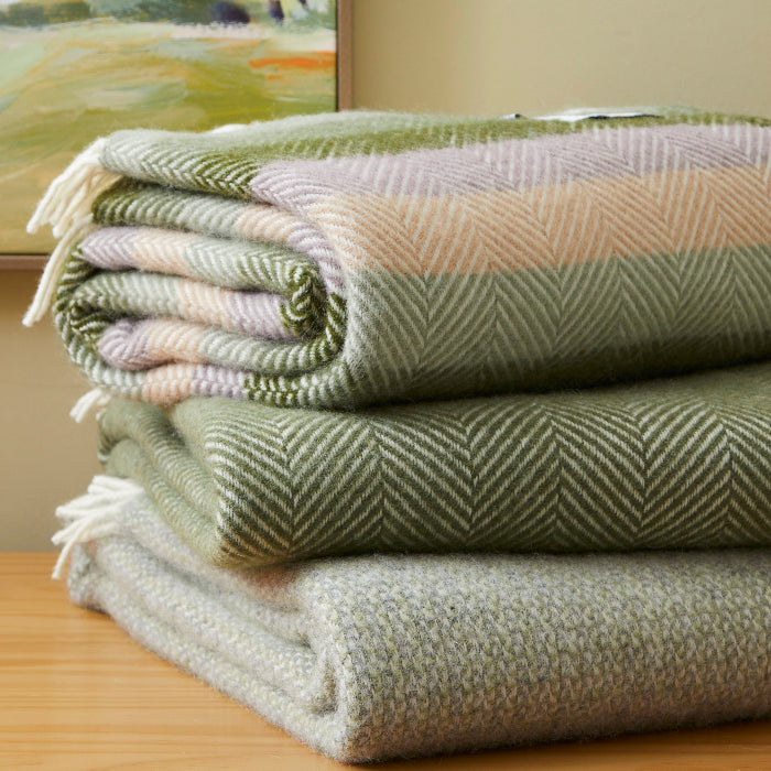 Stack of green wool textured blankets on a wooden surface, in front of a neutral wall with a painting on it in the background