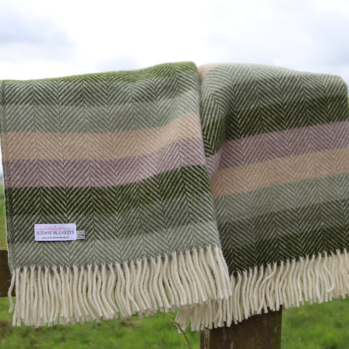 Two multi green toned stripe woollen blankets with fringes displaying a 'Solway Blankets' label, draped over a wooden fence against a scenic background.