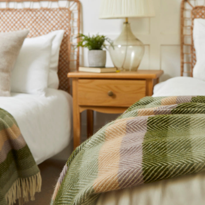 Bedroom with two beds with wicker headboards with green and beige mixed stripe wool blankets draped over the foot of the bed, and a nightstand with lamp.