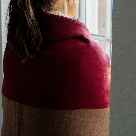 Person wrapped in a burgundy red and tan cashmere throw standing in front of a window.