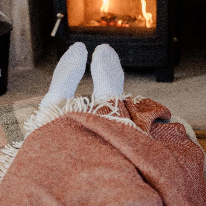 Feet in white socks wrapped in a Brick Herringbone Shetland Wool Blanket by a log burner stove with flames