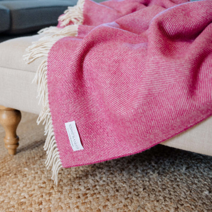 Bright Pink Herringbone Shetland Wool Blanket with a Solway Blankets brand tag draped over a beige ottoman in a living room.