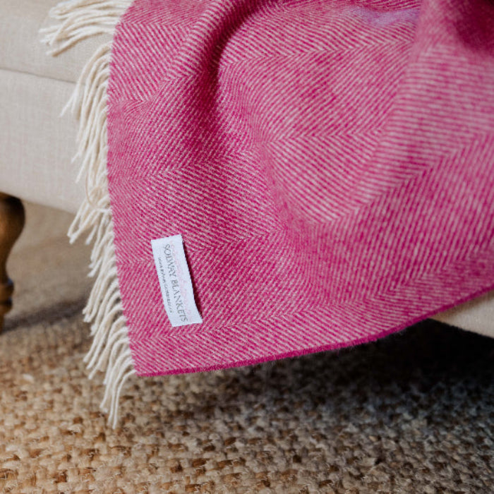 Bright Pink Herringbone Shetland Wool Blanket throw on a beige ottoman with a visible brand label on a carpeted floor.