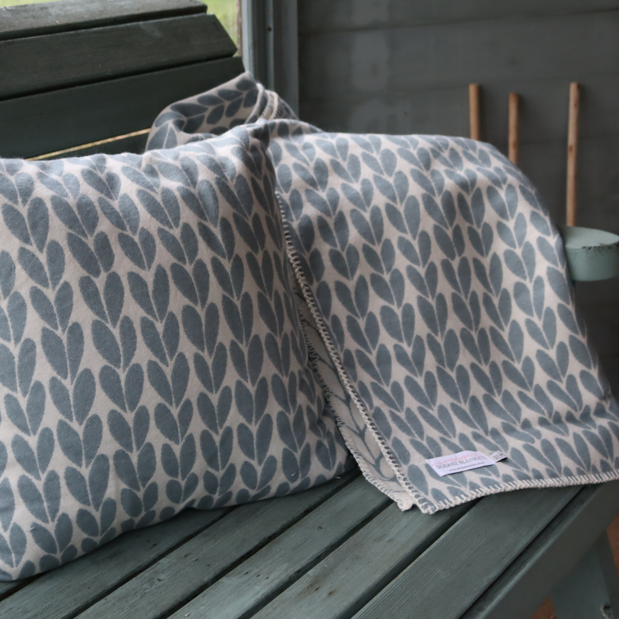 A soft cotton throw cushion and matching blanket with a duck egg blue sycamore leaf pattern on a cream background, draped over a weathered green wooden bench.