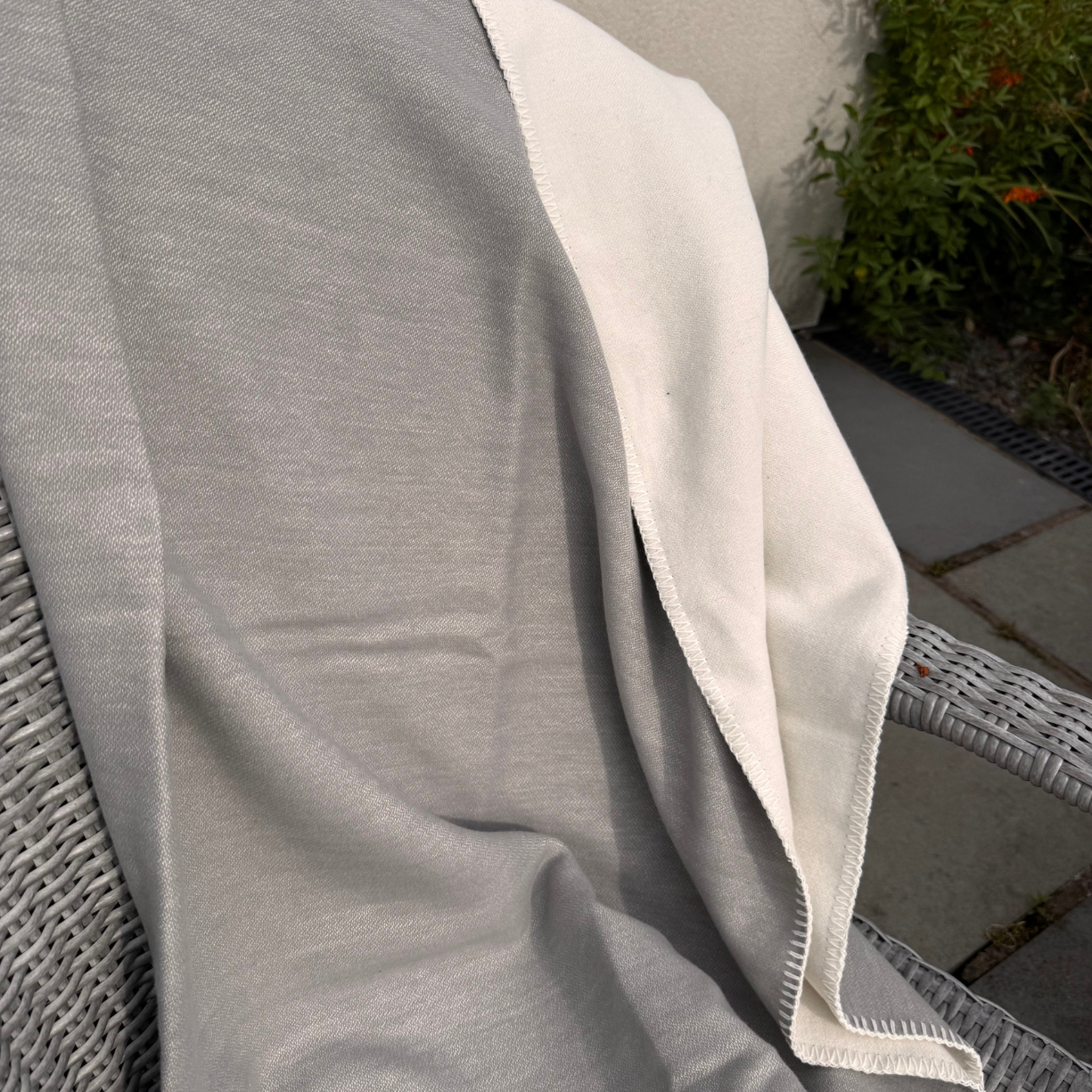 A light grey cotton reversible blanket draped, with the reverse side showing an off-white/cream and a blanket stitched edge, set in an outdoor garden environment.