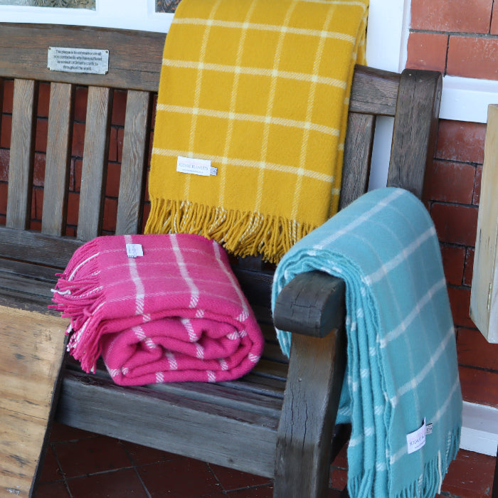 Wooden bench with three colorful chequered wool blankets with colours of yellow, pink and blue draped over it, placed against a brick wall.