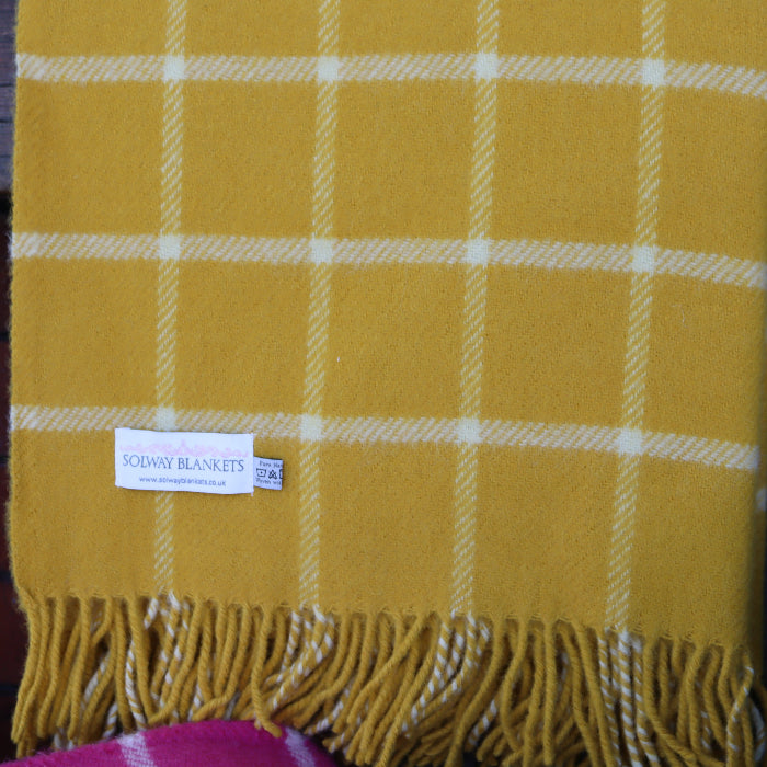 Yellow and white chequered wool blanket with fringes with a 'Solway Blankets' label draped over a wooden bench outdoors