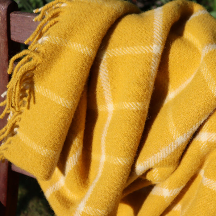 Close up of a yellow and white chequered wool blanket with fringes,  draped over a wooden chair outdoors with a grassy background