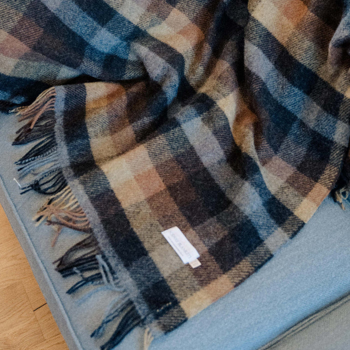 Plaid wool blanket draped over a blue couch featuring colours of blue, navy, tan and chocolate. 