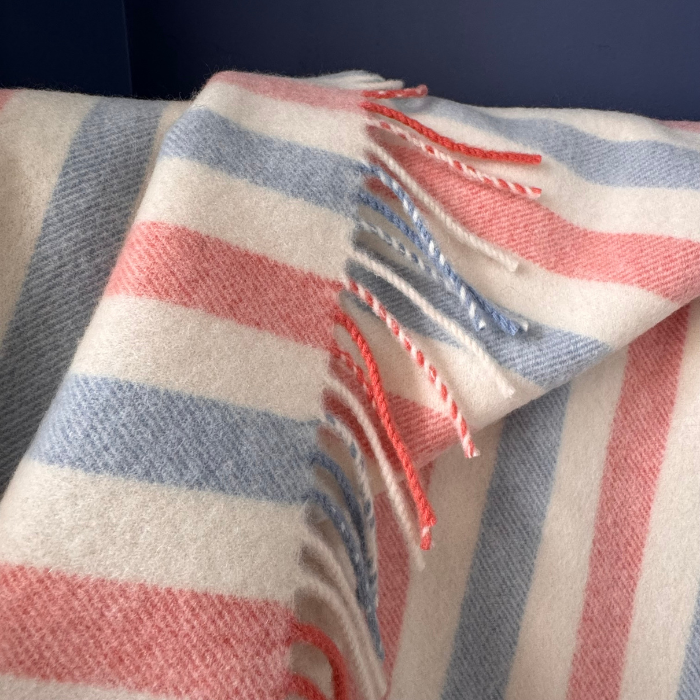 A close-up of a soft cashmere woven baby blanket with stripes in cream, dusty blue and coral pink, finished with fringed edges. Made in Britain