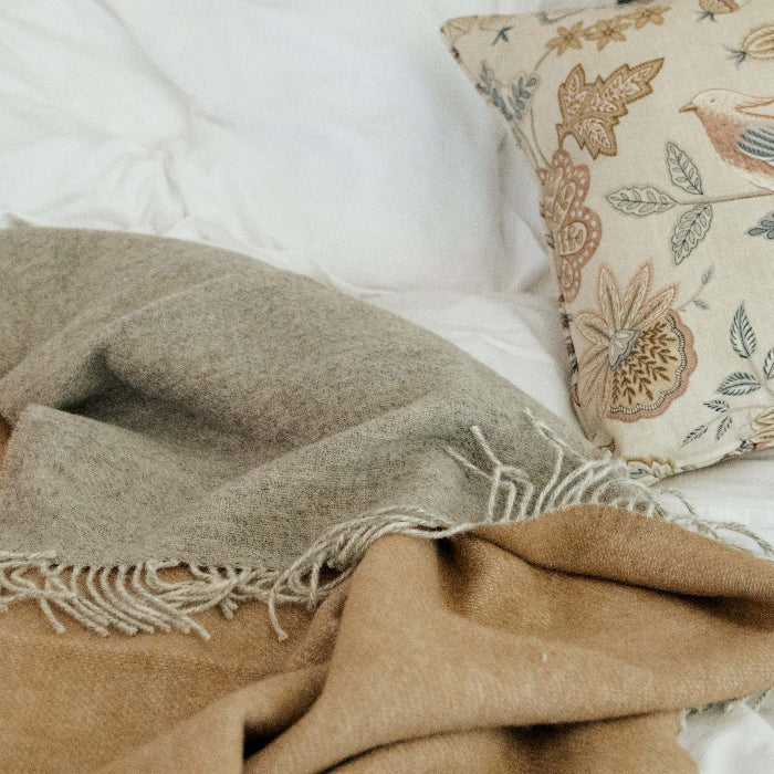 Brown blanket with a soft grey colour on the reverse and a patterned cushion on a white bed