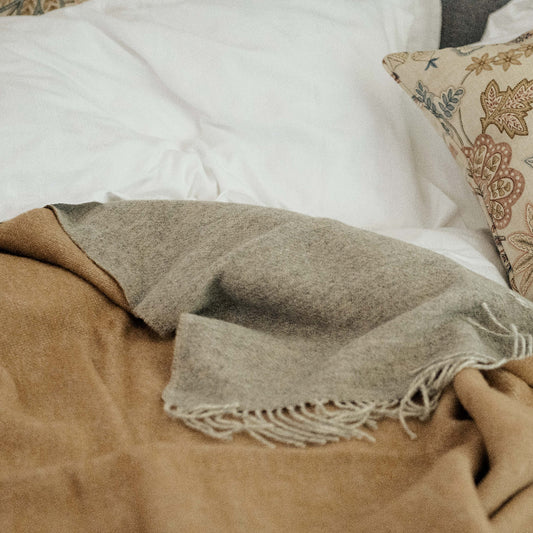 Close up of a bed with white pillows and a patterned cushion, underneath a brown blanket with a soft grey colour on the reverse with fringes. 