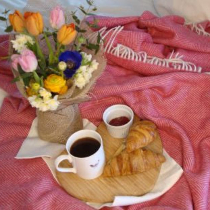 Bouquet of flowers, mugs of coffee, and pastries sitting on top of a pink herringbone patterned blanket throw with white fringe