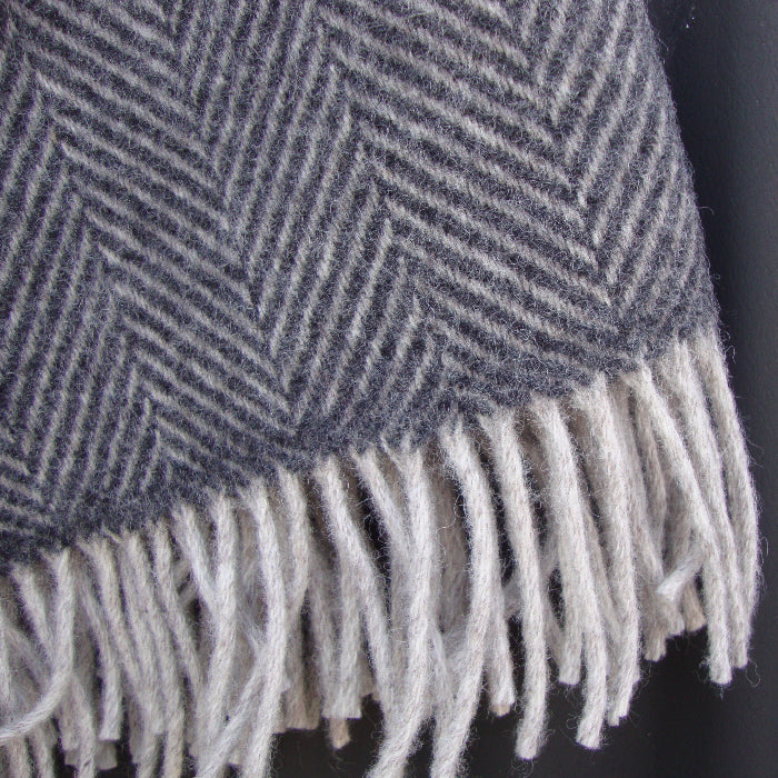 Close up of a charcoal and silver herringbone wool blanket with white fringe pictured outdoors