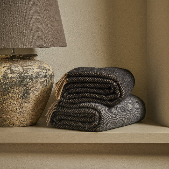 Two folded herringbone style blankets with fringes next to a textured lamp on a shelf against a beige wall.