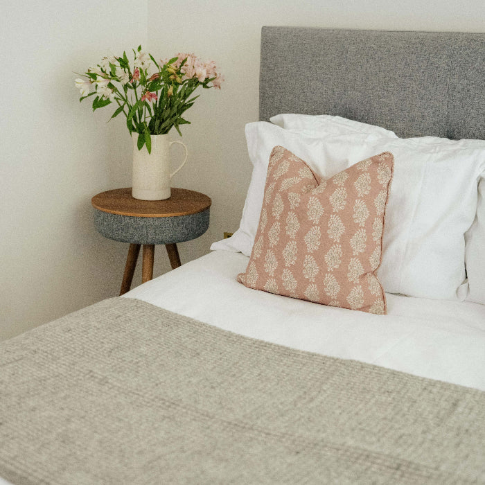 Cloud grey waffle textured wool throw draped on bed with rose coloured cushions and a bedside table with a vase and flowers