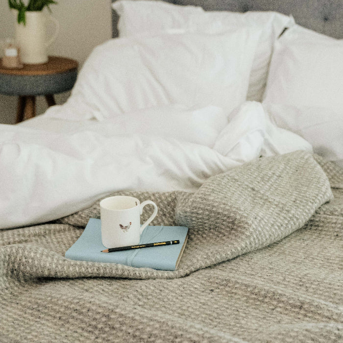 White duvet on a bed with a cloud grey waffle textured wool throw draped on top featuring a mug, notebook, and a pencil 