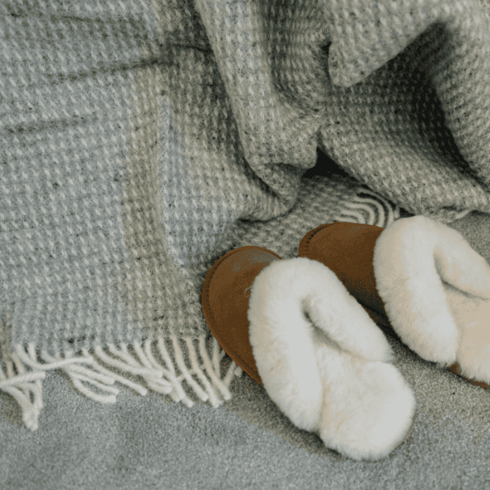 Cloud grey waffle textured wool throw with cream fringe draped beside soft sheepskin slippers on a bedroom carpet – cosy, calm and beautifully layered.
