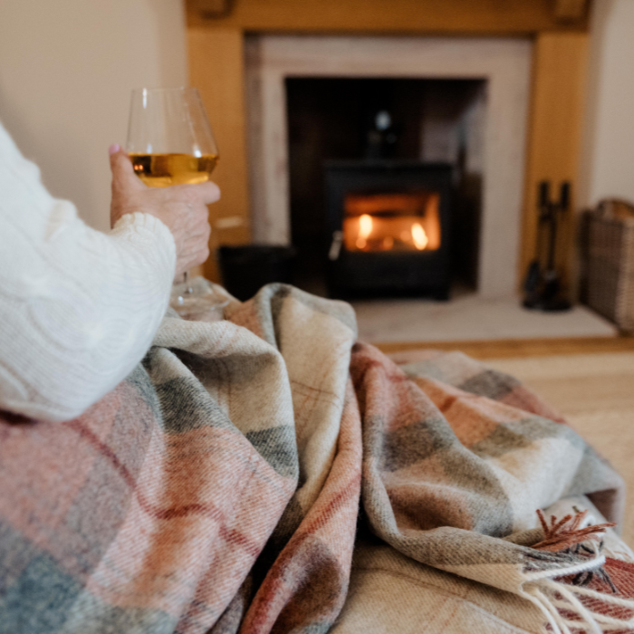 Person wrapped in the Coral Skye Check Shetland Wool Throw plaid blanket holding a glass of wine in front of a fireplace.