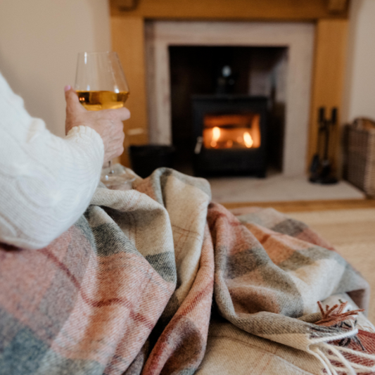 Person wrapped in the Coral Skye Check Shetland Wool Throw plaid blanket holding a glass of wine in front of a fireplace.