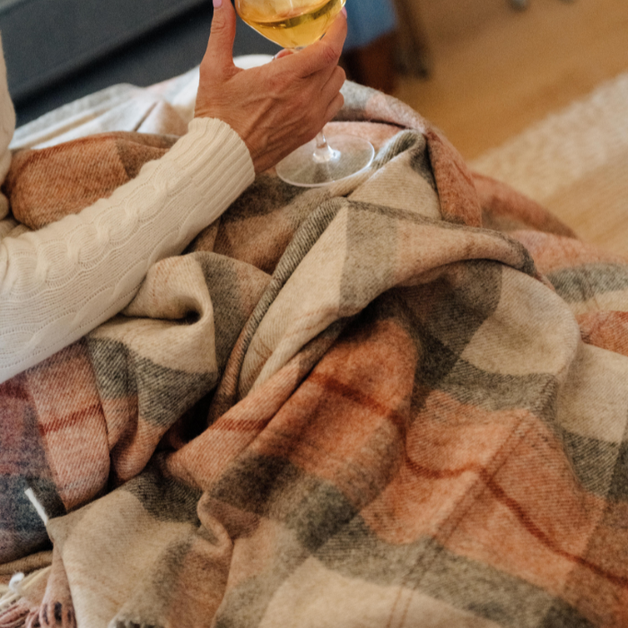 Person wrapped in the Coral Skye Check Shetland Wool Throw plaid blanket holding a glass of wine indoors.