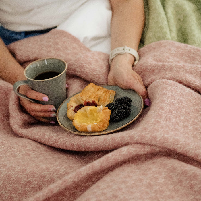 Person holding a cup of coffee and a plate of pastries and blackberries, wrapped in a soft pink wool throw 