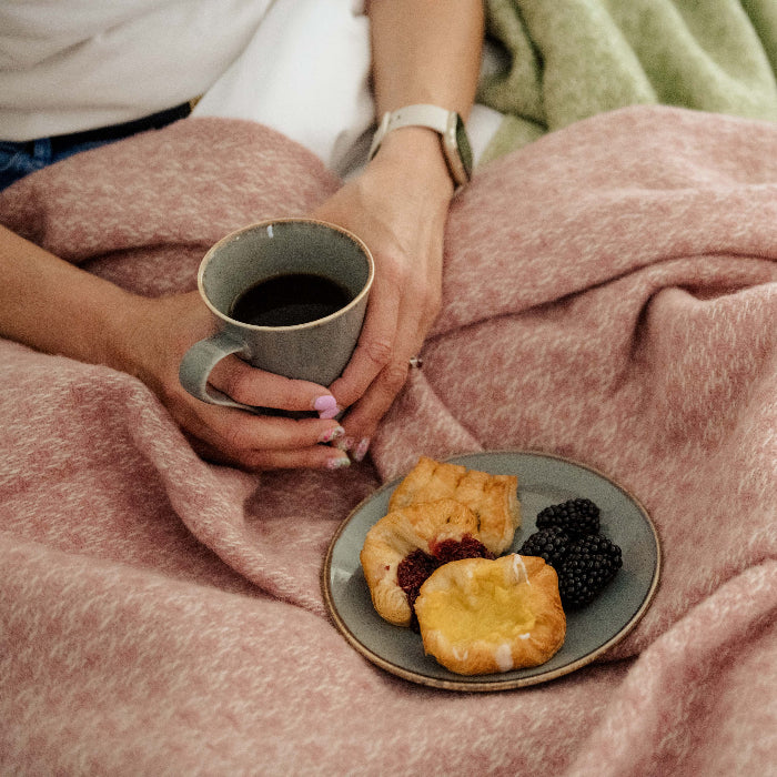 Person holding a mug of coffee with a plate of pastries and berries under a soft pink woollen blanket.