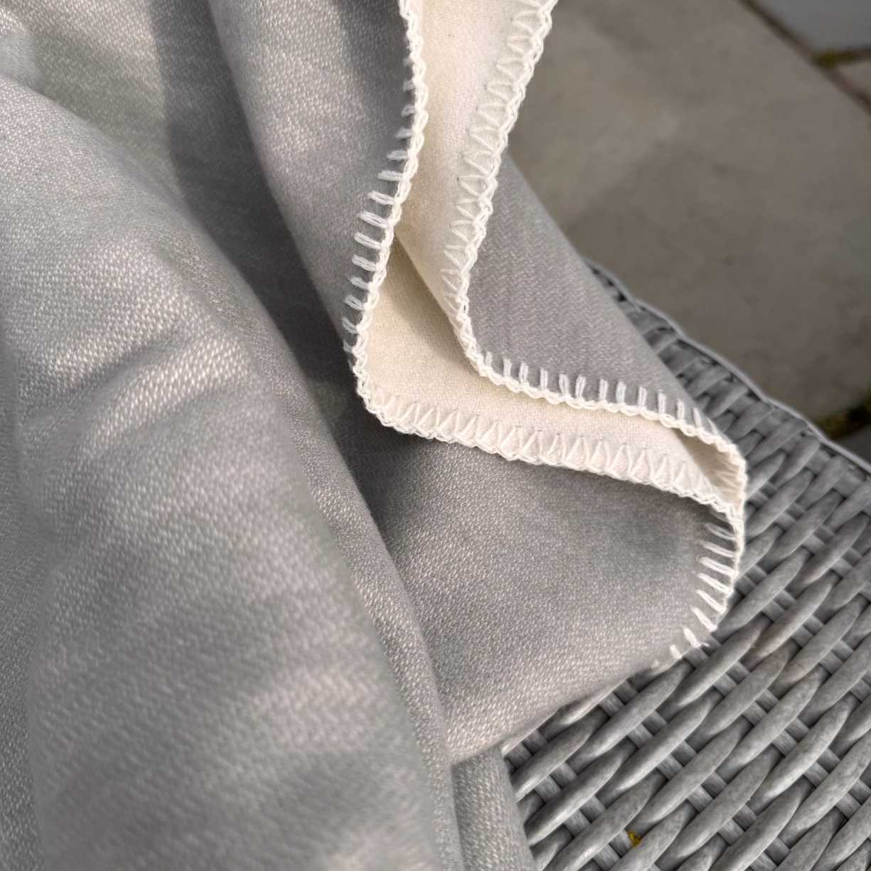 A close-up of a light grey cotton throw blanket draped over a grey wicker chair, showing a contrasting off-white stitched edge and reverse side