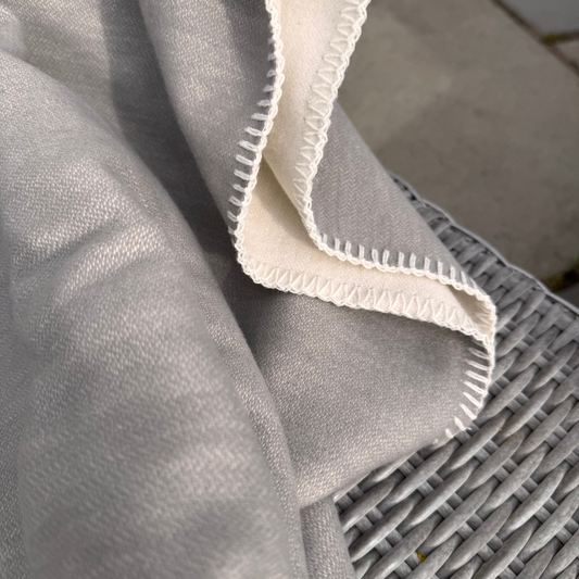 A close-up of a light grey cotton throw blanket draped over a grey wicker chair, showing a contrasting off-white stitched edge and reverse side
