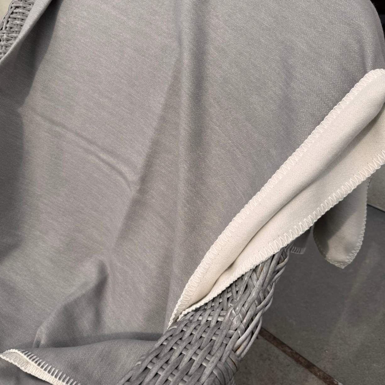 A close-up of a light grey plain cotton blanket draped over a gray woven wicker chair. The fabric has a soft texture and a contrasting off-white/cream stitched border and white reverse