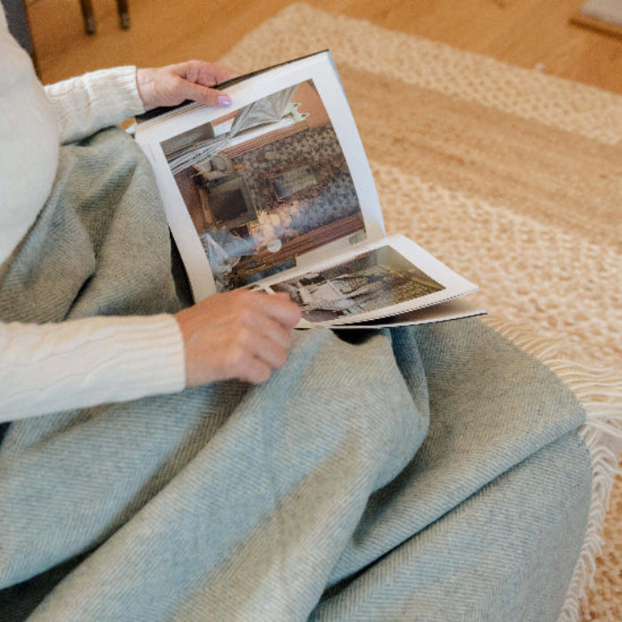 Lady reading a coffee table book with the Duck Egg Blue Herringbone Shetland Wool Blanket over her knee