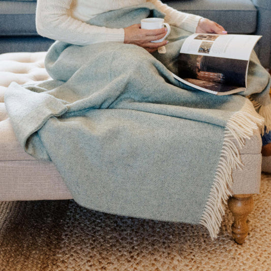 Person sitting on a couch with a Duck Egg Blue Herringbone Shetland Wool throw, holding a mug of tea and reading a magazine.