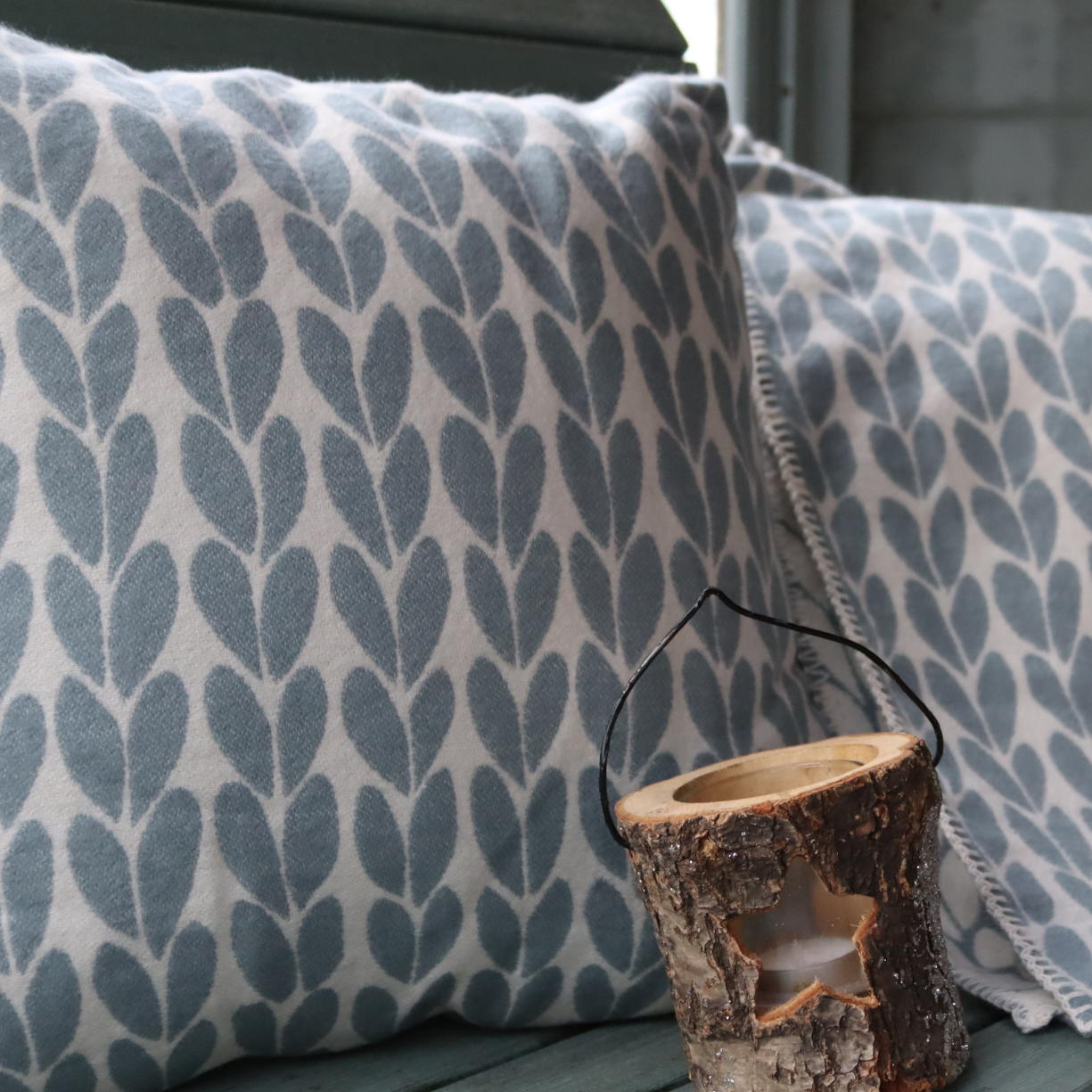 A decorative pure cotton throw pillow with a duck egg blue leaf pattern on a white background, styled on a dark green bench, with a matching throw next to a rustic wooden candle holder.