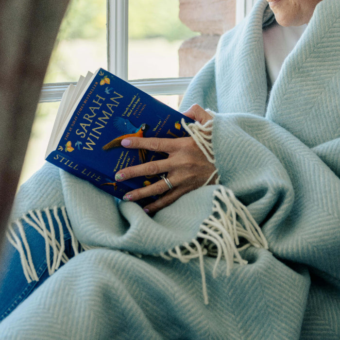 Person wrapped up in a duck egg fishbone wool blanket holding a book titled 'Still Life' by Sarah Winman.