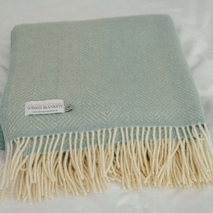 Duck egg coloured fishbone style wool blanket with fringe displaying a Solway Blankets label, folded on a white soft surface. 