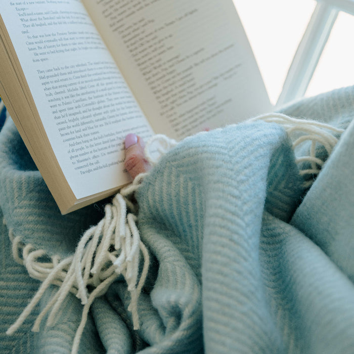 Person wrapped in a duck egg coloured fishbone wool blanket reading a book