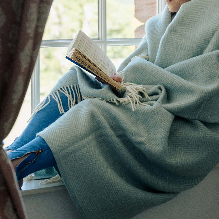 Person sitting by a window reading a book, wrapped up in a duck egg coloured fishbone style woolen blanket