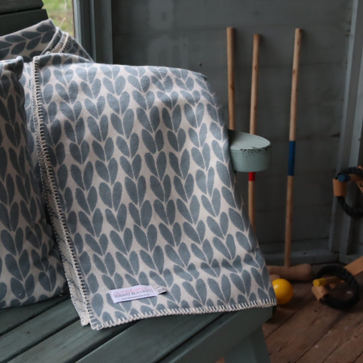 Duck egg blue, sycamore leaf design on a cotton British made throw, draped over a garden bench arm with croquet garden game set in the background.