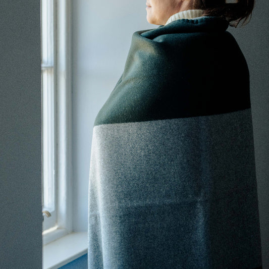 Person wrapped in a bottle green and soft grey cashmere blanket throw looking out of a window.