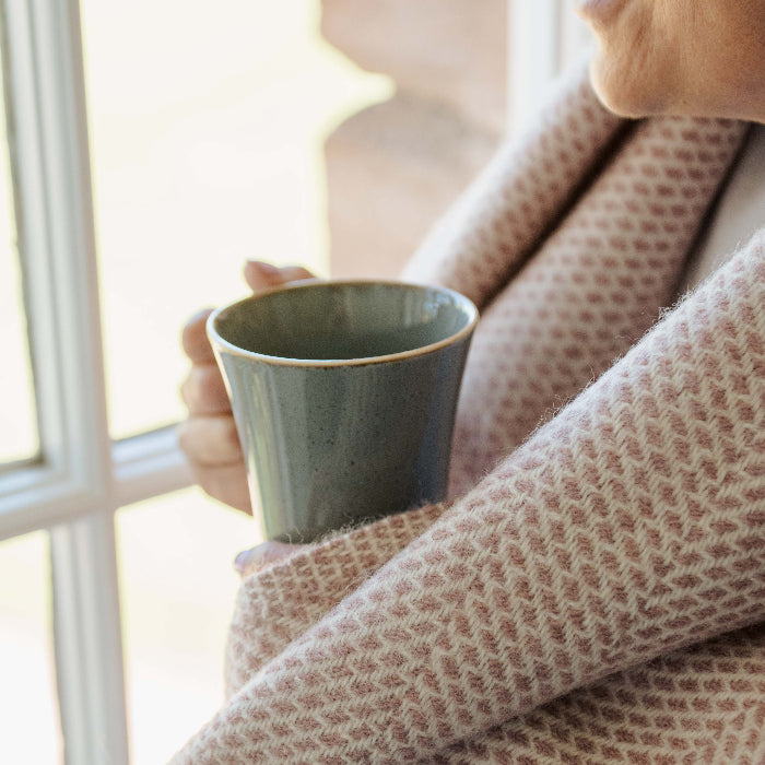 Person wrapped in a cosy dusky pink blanket holding a mug by a window