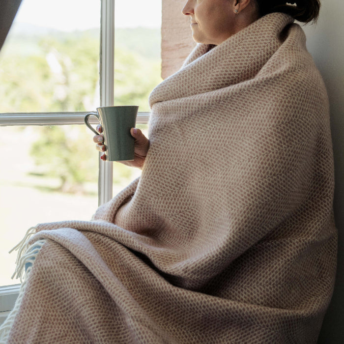 Person wrapped in a dusky pink beehive pattern woollen throw holding a mug looking out a window