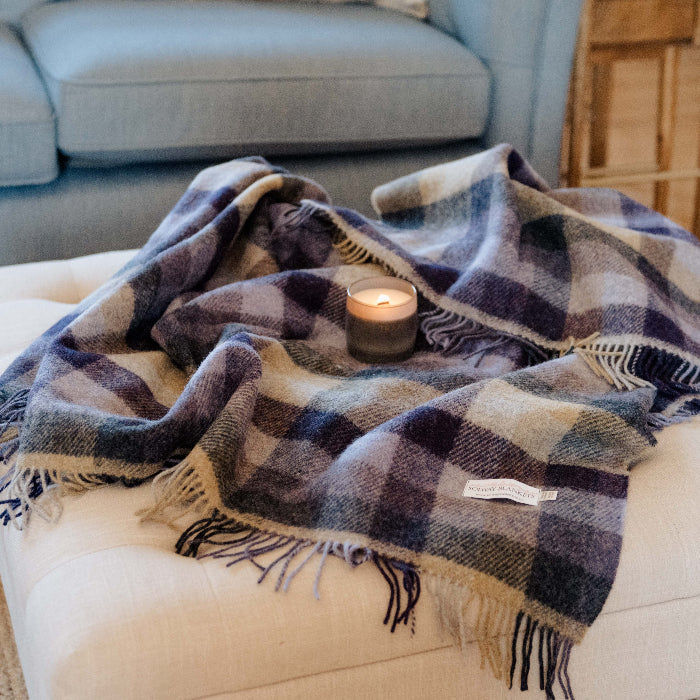Lilac, grey and beige plaid wool blanket draped over the top of a pouffe, with a lit candle in a jar on top, in a living room setting. 