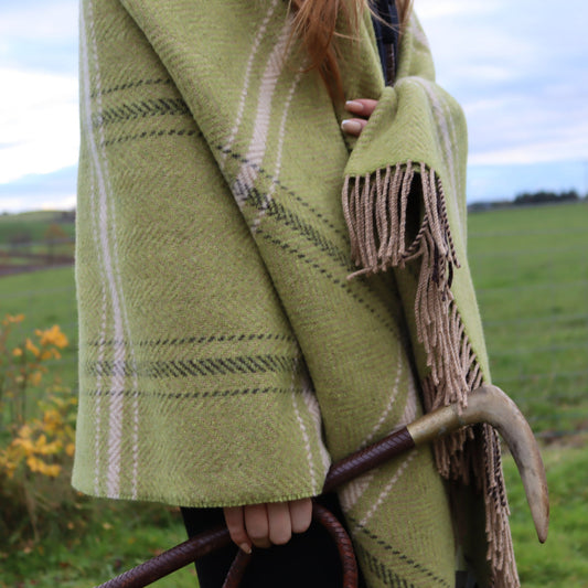Person wearing a emerald green plaid throw with fringe, standing in a field.