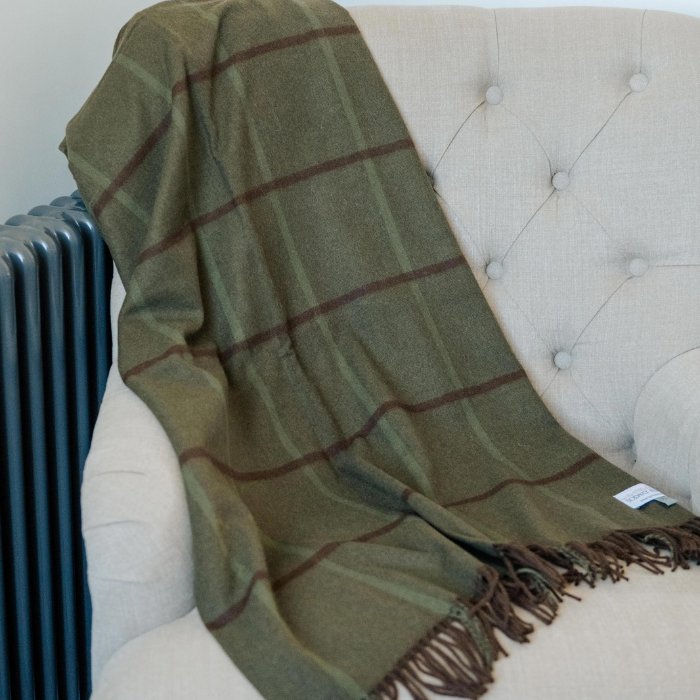 A large olive green plaid cashmere throw blanket with dark brown and green large check and fringe, draped over a cream tufted armchair.