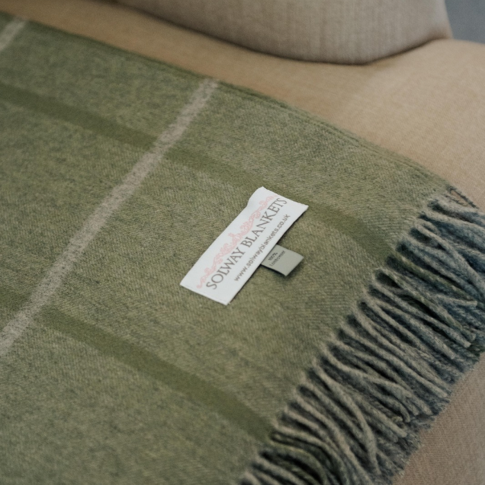 Close-up of fine sage green cashmere woven throw with pale windowpane check. Showing delicate calming green tones with a pale grey and meadow green over check design, finished with a beautiful twisted fringe and brand label 'Solway Blankets'
